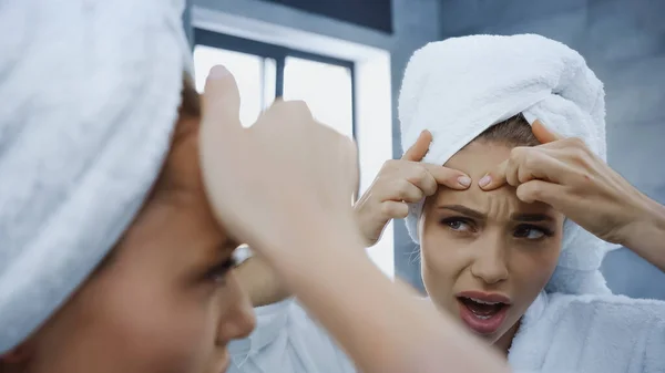 Displeased young woman squeezing pimple and looking at mirror in bathroom — Stock Photo