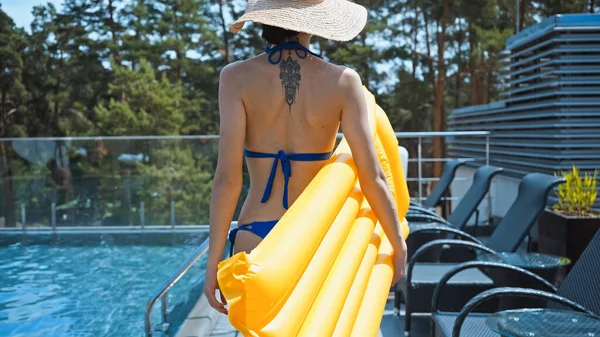 Back view of tattooed woman in straw hat holding inflatable mattress and walking near pool — Stock Photo