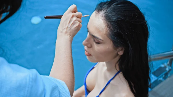 Visão de alto ângulo do artista de maquiagem aplicando pó facial com escova cosmética na mulher morena perto da piscina — Fotografia de Stock