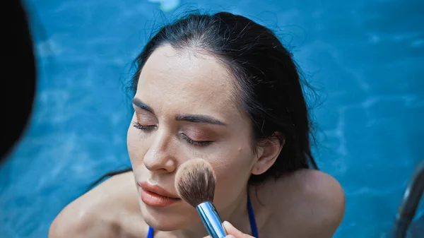 Artista de maquiagem aplicando pó facial com escova cosmética na mulher morena perto da piscina — Fotografia de Stock