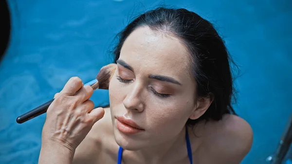 Maquillaje artista aplicar polvo facial con cepillo cosmético en la mujer joven cerca de la piscina - foto de stock