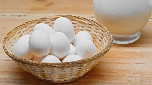 Panier en osier avec des œufs blancs près du bocal en verre avec du lait frais sur la surface en bois — Photo de stock