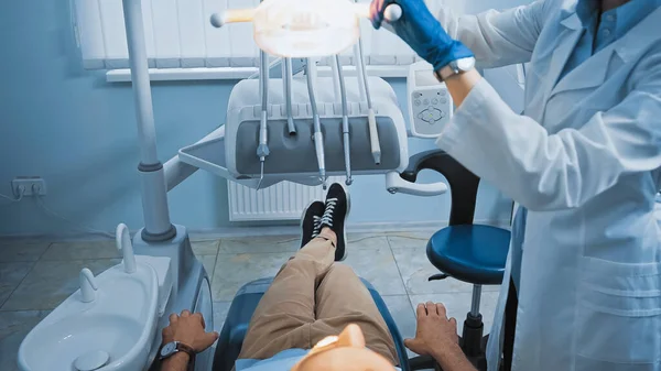Vista ritagliata del dentista lampada di regolazione sopra paziente seduto sulla sedia medica — Foto stock