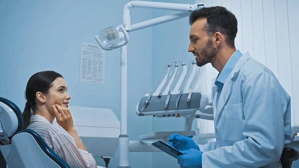 Vista lateral do dentista com área de transferência perto da mulher apontando para a bochecha na cadeira médica — Fotografia de Stock