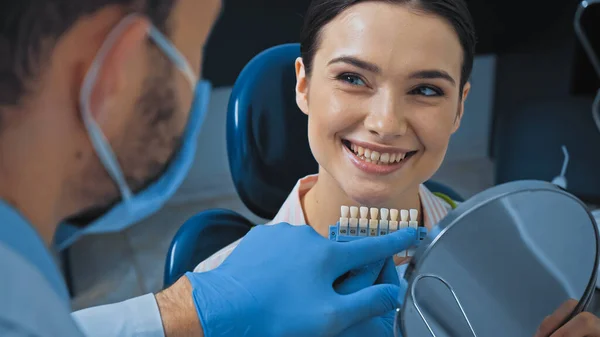 Dentist pointing with finger at teeth color palette near smiling woman, blurred foreground — Stock Photo