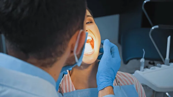 Dentiste en masque médical examinant les dents de la femme en clinique, avant-plan flou — Photo de stock