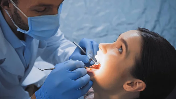 Estomatólogo en máscara médica examinar los dientes del paciente con instrumentos dentales - foto de stock
