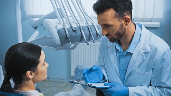 Dentista segurando prancheta enquanto conversa com o paciente perto de instrumentos dentários — Fotografia de Stock