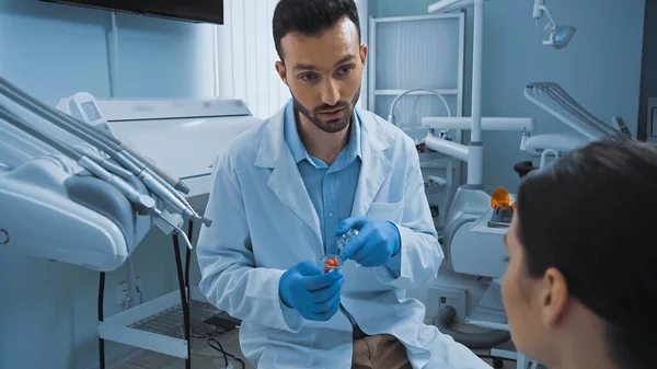 Dentista apontando para o modelo de dentes enquanto conversa com o paciente na clínica, foreground borrado — Fotografia de Stock