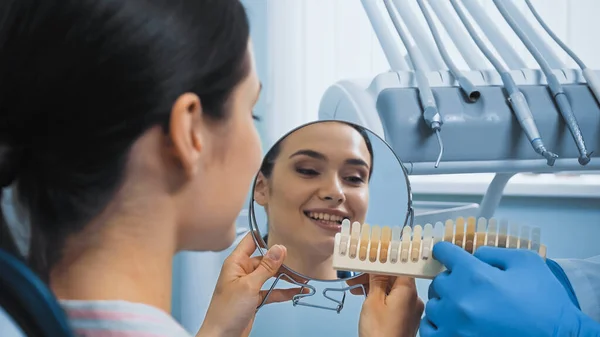 Dentist showing teeth color palette to woman holding mirror, blurred foreground — Stock Photo