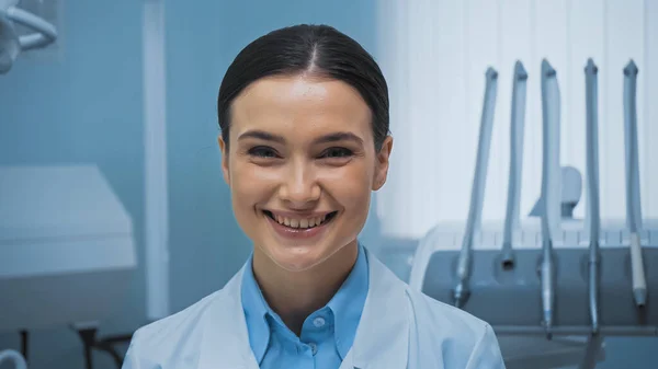 Dentista alegre sorrindo para a câmera perto de equipamentos odontológicos em fundo embaçado — Fotografia de Stock