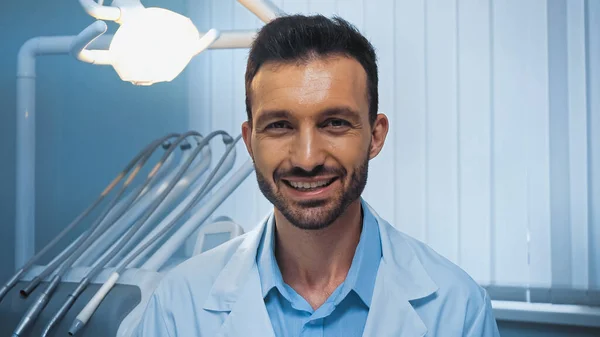 Счастливый стоматолог смотрит в камеру рядом с стоматологическим оборудованием и лампой на размытом фоне — стоковое фото