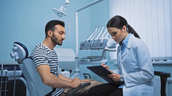 Bonito dentista escrever prescrição perto de homem sentado na cadeira médica — Fotografia de Stock