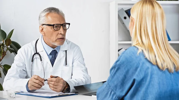 Mature doctor holding pen while talking to woman on blurred foreground — Stock Photo