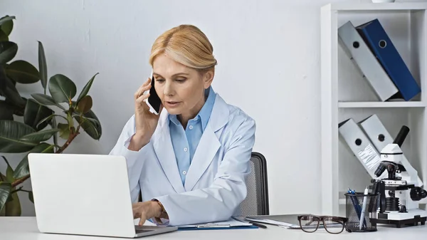 Mature doctor talking on smartphone near laptop at workplace — Stock Photo