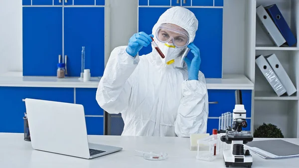Scientist in hazmat suit talking on smartphone while holding test tube with blood sample — Stock Photo