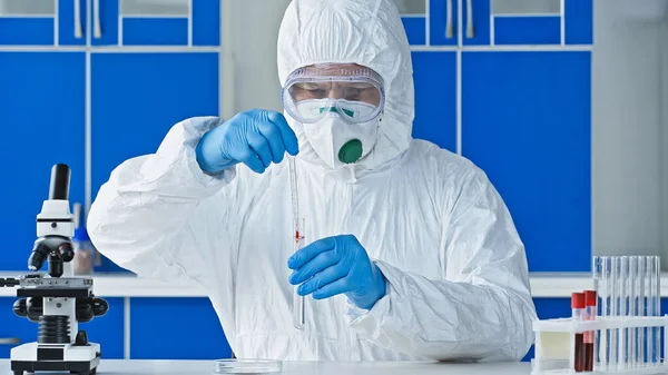 Wissenschaftler im Schutzanzug hält Reagenzglas und Tropfer während er in der Nähe des Mikroskops arbeitet — Stockfoto