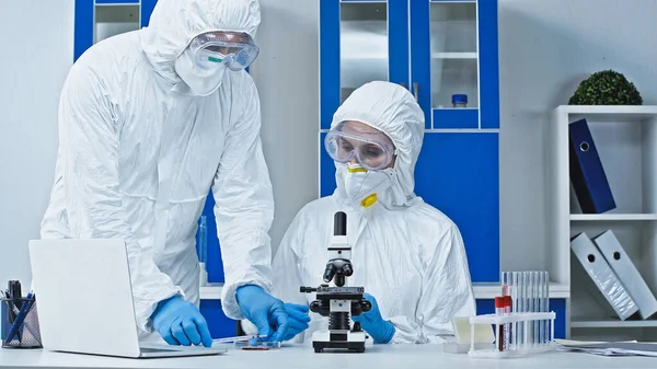 Scienziato in tuta antirischio che mostra il campione di sangue al collega vicino al microscopio — Foto stock