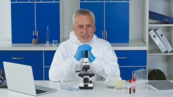 Lächelnder Wissenschaftler im Warnanzug, der in die Kamera neben Mikroskop und Laptop blickt — Stockfoto