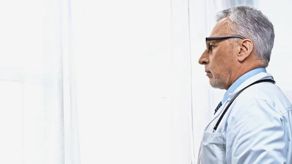 Seitenansicht eines reifen Arztes, der in der Nähe des Fensters im Krankenhaus wegschaut — Stockfoto