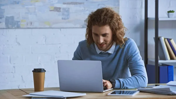 Uomo d'affari riccio digitando sul computer portatile vicino al caffè per andare sul posto di lavoro — Foto stock