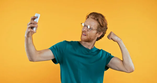 Redhead man in glasses showing muscle while taking selfie isolated on yellow — Stock Photo