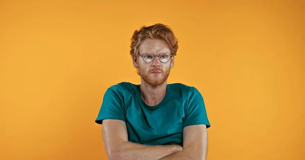 Offended redhead man in glasses standing with crossed arms isolated on yellow — Stock Photo