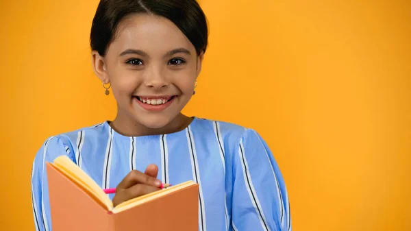 Niño alegre sosteniendo lápiz y cuaderno aislado en amarillo - foto de stock