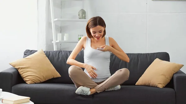 Счастливая беременная женщина трогает живот, отправляя голосовое сообщение на мобильный телефон — стоковое фото