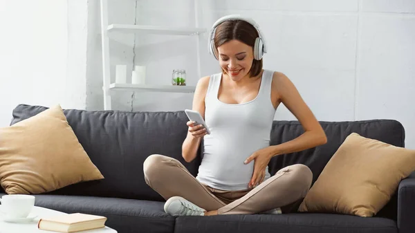 Mulher grávida feliz sentado no sofá, ouvindo música e conversando no smartphone — Fotografia de Stock