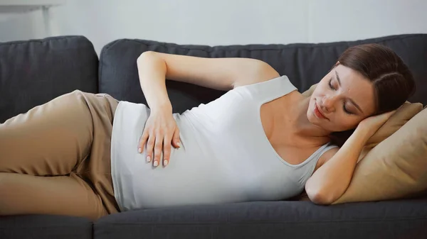 Pretty, pregnant woman touching belly while resting on sofa at home — Stock Photo