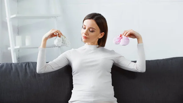 Young pregnant woman holding baby booties while sitting on couch — Stock Photo