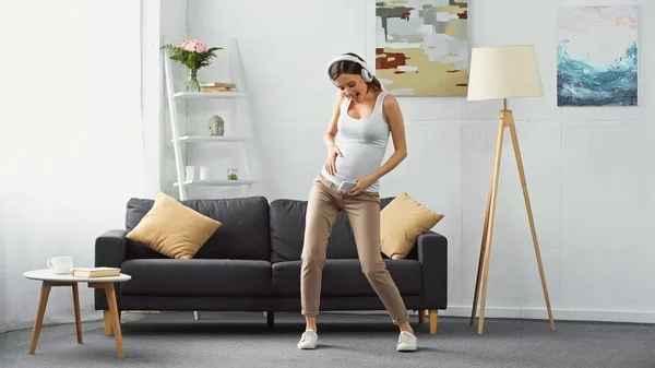 Vista a tutta lunghezza della donna incinta eccitata che tiene smartphone mentre balla in cuffia — Foto stock