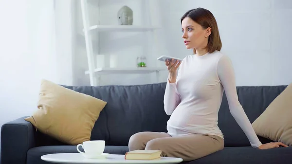 Donna incinta che invia messaggi vocali su smartphone vicino alla tazza e prenota sul tavolino — Foto stock