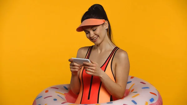 Giovane donna allegra in costume da bagno e anello gonfiabile utilizzando smartphone isolato su giallo — Foto stock