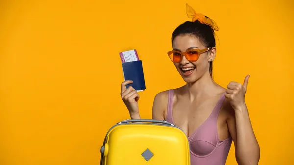 Mujer sonriente sosteniendo pasaporte y billete de avión cerca de la maleta y mostrando el pulgar hacia arriba aislado en amarillo - foto de stock