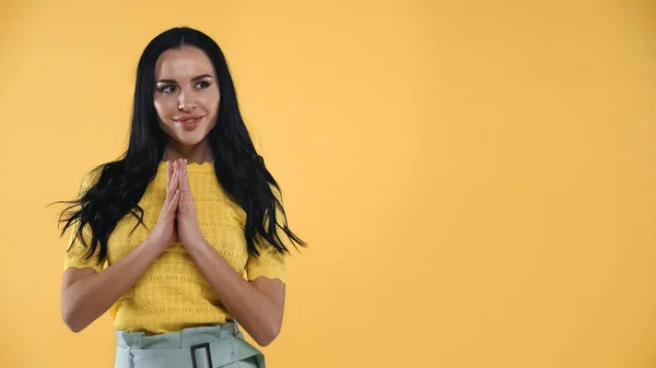 Positive brunette woman standing with praying hands isolated on yellow — Stock Photo