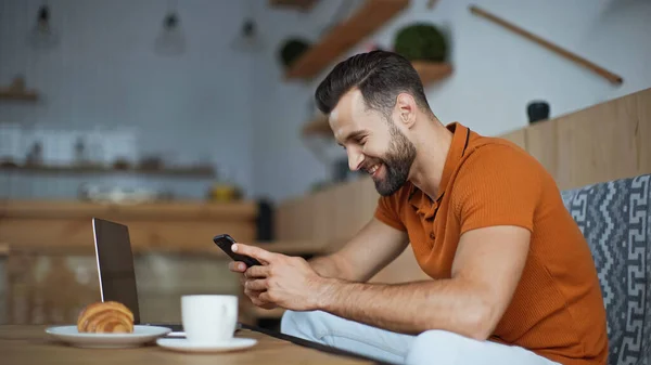 Mensajería freelancer feliz en el teléfono inteligente cerca del ordenador portátil y el desayuno en la cafetería - foto de stock