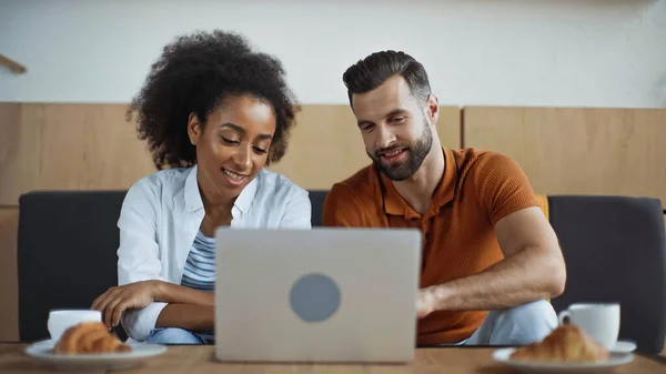 Alegres freelancers interracial mirando el ordenador portátil y el desayuno en la cafetería - foto de stock