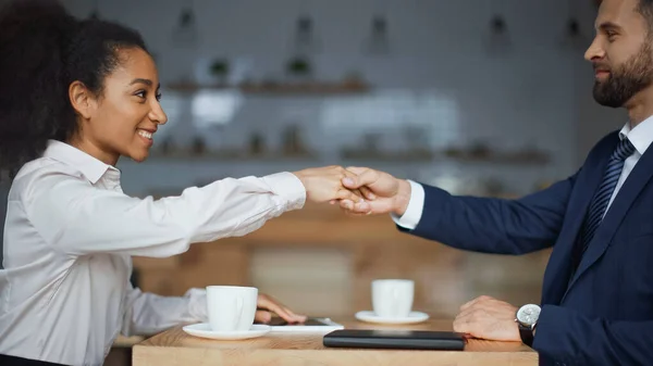 Side view of happy interracial business partners shaking hands during morning meeting in cafe — Stock Photo