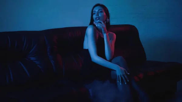 Iluminación roja en la cara de la mujer morena pensativa sentado en el sofá negro en azul - foto de stock