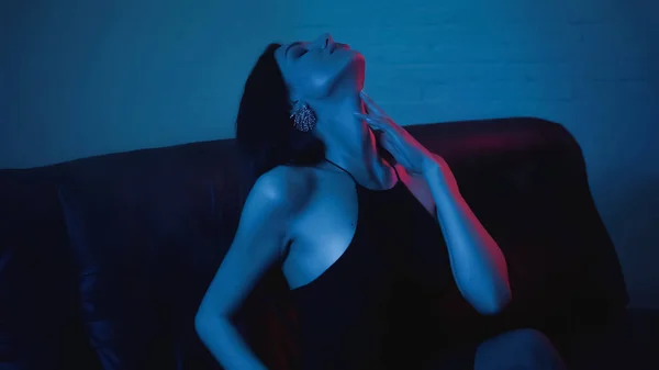 Illuminazione rossa sulla seducente donna bruna che tocca il collo sul blu con il fumo — Foto stock