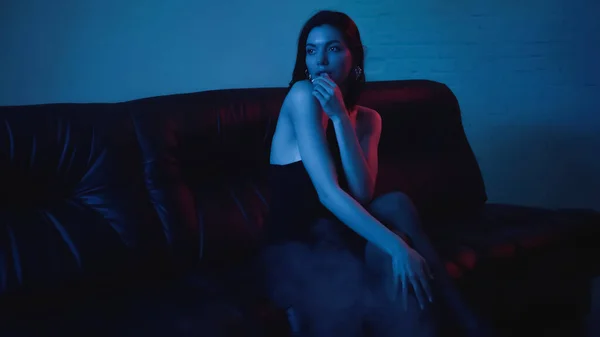 Éclairage rouge sur séduisante brune femme sur canapé noir sur bleu avec fumée — Photo de stock