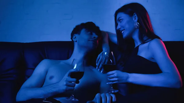 Coppia sexy che tiene bicchieri di vino e sorridente mentre si guarda l'un l'altro sul blu — Foto stock