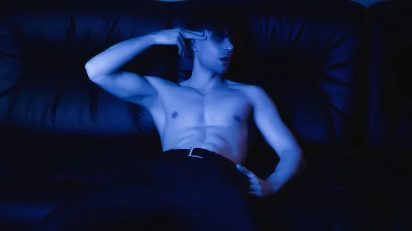 Illuminazione blu su uomo senza maglietta in posa e gesticolando mentre riposava sul divano nero — Foto stock