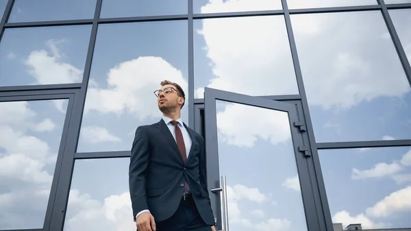 Vista de ángulo bajo del hombre de negocios en traje y gafas que abren la puerta mientras camina fuera - foto de stock