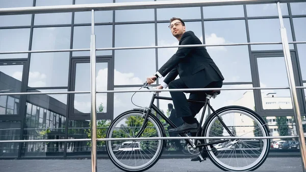 Full length of businessman in suit riding bicycle near building with glass facade — Stock Photo