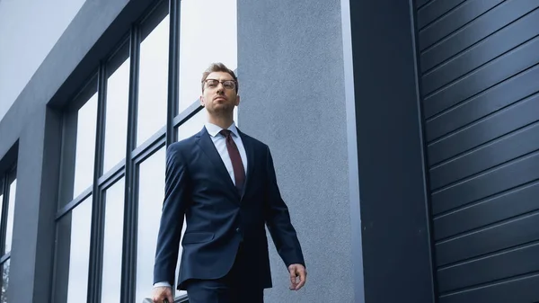 Hombre de negocios confiado en traje y gafas caminando cerca del edificio - foto de stock