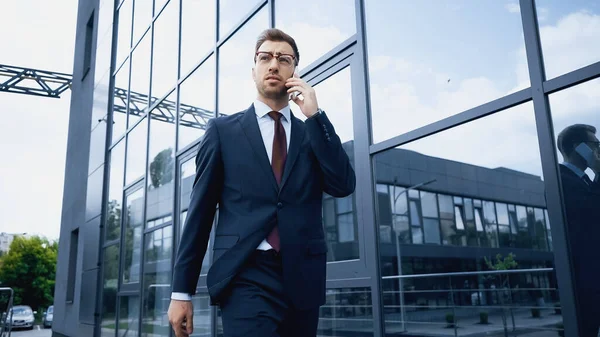 Confident businessman in glasses and suit talking on smartphone while walking near building — Stock Photo