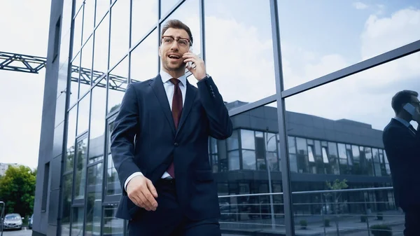 Businessman in glasses and suit talking on smartphone while walking near building — Stock Photo